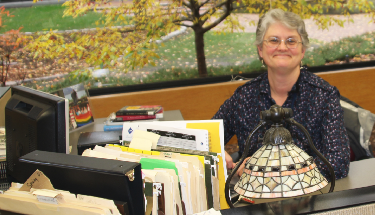 Vicki Janisch-Tri smiles for a picture while working at her desk. “I like the chance to get to know the students. I enjoy working with [Upper School librarian] Ms. Brooks and making sure that the collection is in good shape for people to come and find what they need. I just like seeing how this school works, [and] the daily lives of students,” Janisch-Tri said.