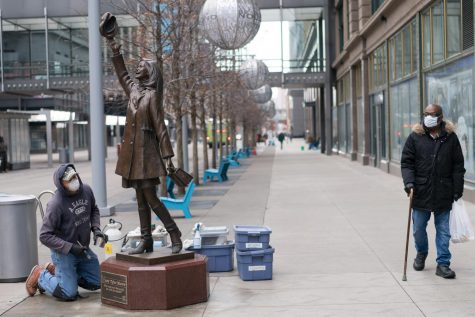 Two people wearing face masks, a worker cleaning the Mary Tyler Moore statue and someone passing by, glance at each other in Minneapolis, Minnesota.