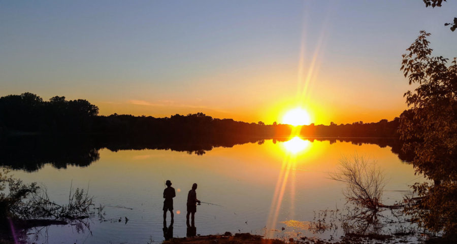 Two men fish on the clear glasslike lake with the sun setting in the distance.