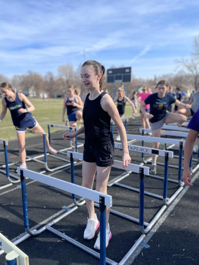 SMILING WHILE HURDLING. Freshman Hazel McCarthy wears a gleaming smile while completing a hurdle warmup. McCarthy moved on to sprinting drills following the warmup and completed it with more enthusiasm. “This is my first year doing high school track and I think it is really fun and challenging. Track practice has given me the opportunity to try a bunch of new things that I would not have normally done,” said McCarthy.