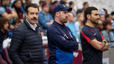 THERAPEUTIC TRIO. Fan favorites Ted Lasso, Coach Beard and Roy Kent stand on the sidelines of the pitch. 