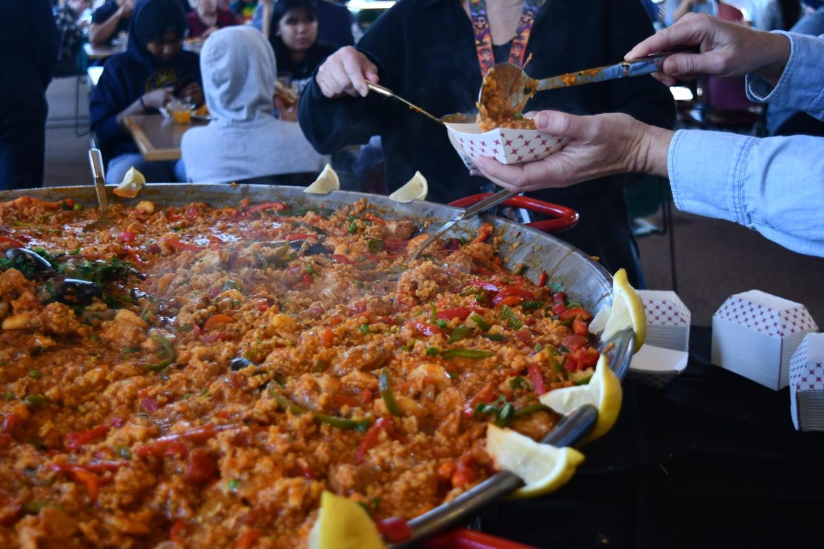 COMMUNAL SERVING. Upper school faculty members serve themselves paella in paper food trays. The display, one large pan of steaming hot paella, was located in the seating area of the dining hall. “We try to … do more stuff on display. So this is kind of a chef on display,” head chef Tom Schiller said.