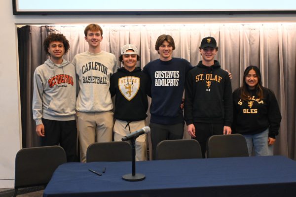 NEW THREADS. Senior athletes pose at the signing ceremony before school on Apr. 17 in their respective college merch. From left to right, Ethan Carter will be playing basketball, Tysen Hayes will be playing basketball, Griffin Schwab-Mahoney will be playing baseball, Cooper Olsen will play soccer, Will Black will play golf and Naomi Kempcke will play basketball. 