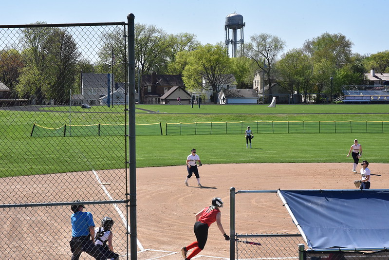TAKEOFF: Redhawks runner swings, drops her bat, and takes off for first base. Breck outfielders and umpire look into the sky to track the ball for an attempted catch. 
