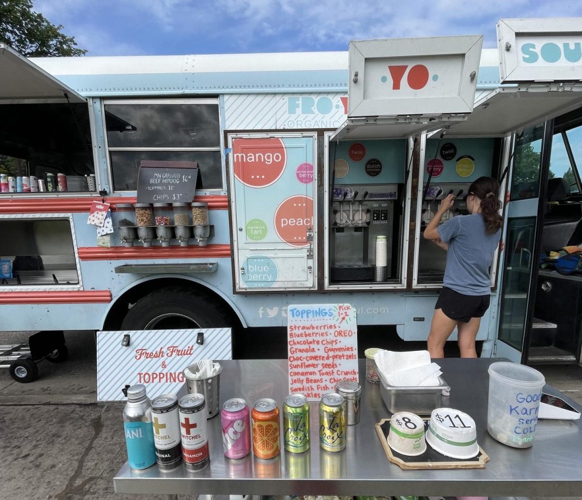 FIRST THOUGHTS. The front of the fro-yo truck is pictured with fro-yo dispensers on the right of the truck and toppings on the left. There is also a table with beverages and pricing info.
