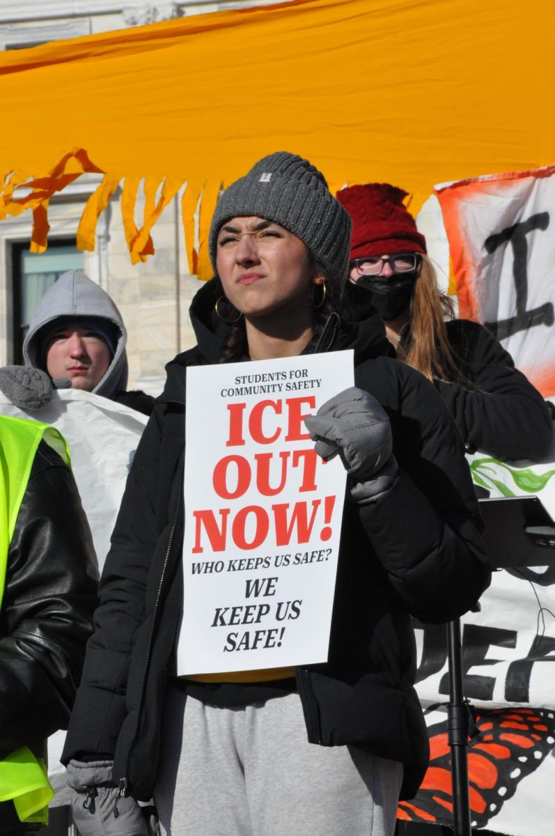 STAND STRONG. A student organizer stands in front of the crowd holding a sign that reads "ICE out now," in bright red letters. 