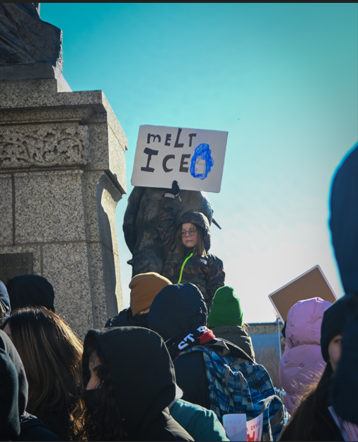 SITTING TALL. A young protester perches on a statue above the crowd, holding a sign with the message, "Melt ICE."